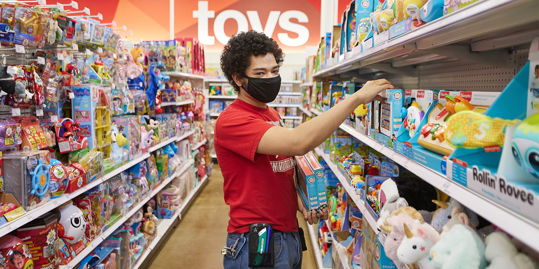 "A Target team member in red shirt and black mask reaches to stock more toys on a shelf."